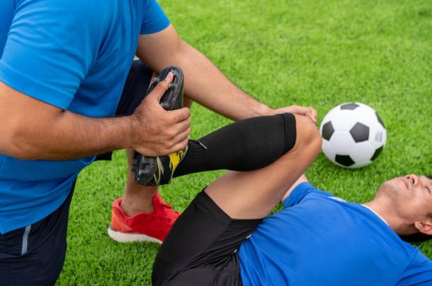 Low Section Of Male Physical Therapist Stretching Soccer Player Leg On Field